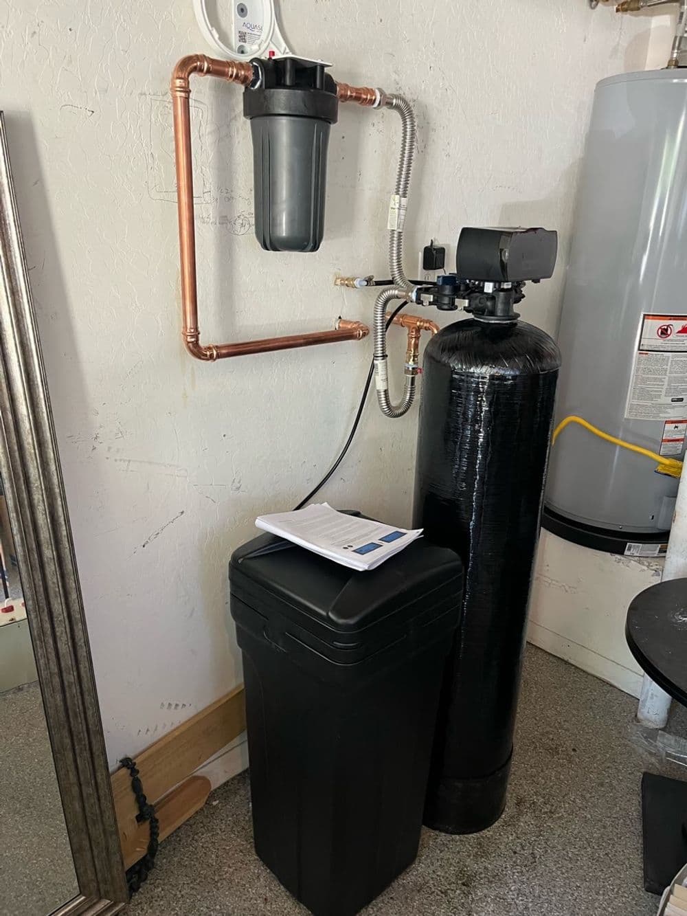 Water filtration system with clear filter housing, black tank, and plumbing connections in garage.