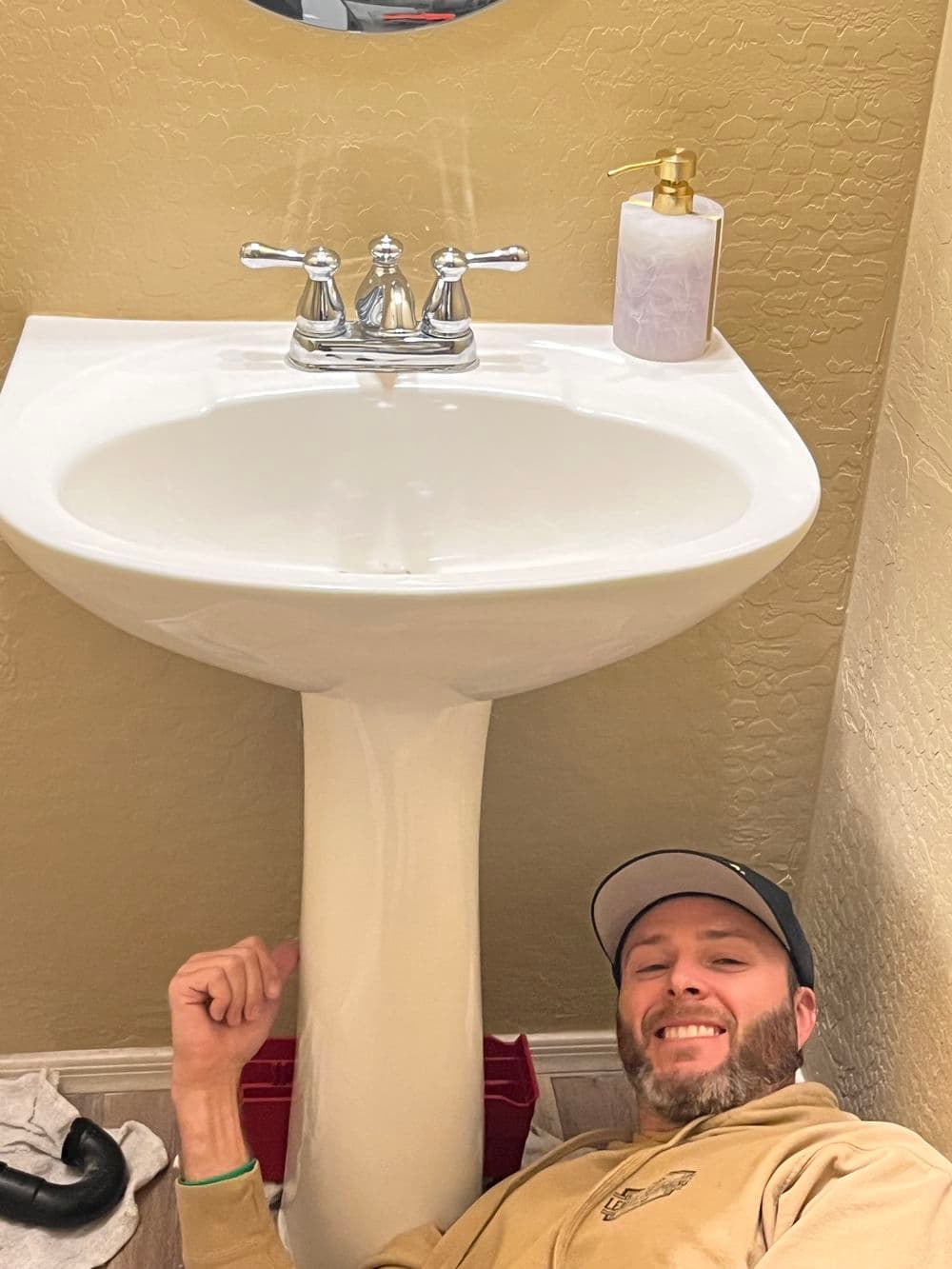 Man smiling while working on a white bathroom sink, with soap dispenser and towels nearby.