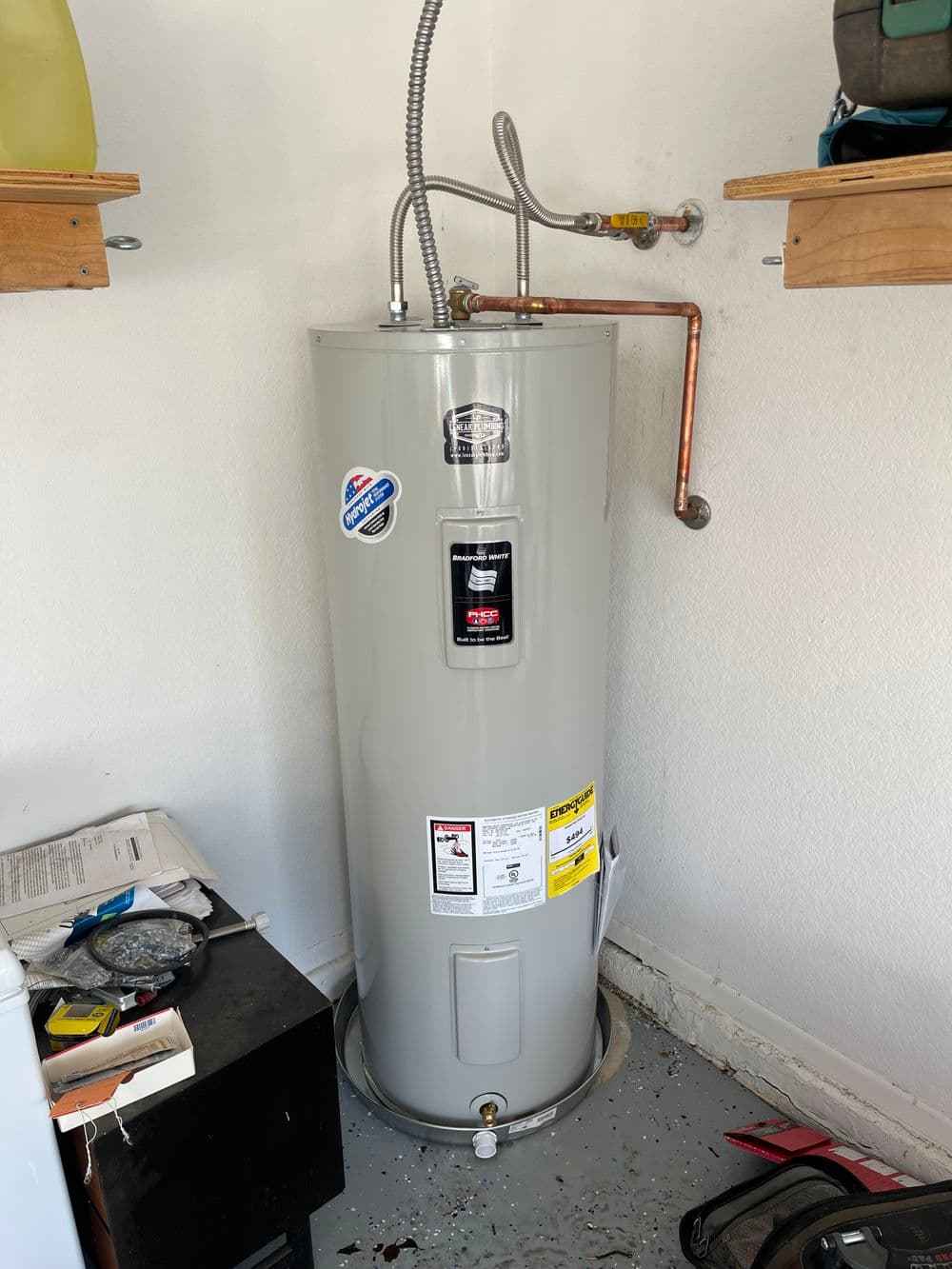 Gray water heater in a garage with copper pipes and shelving above.