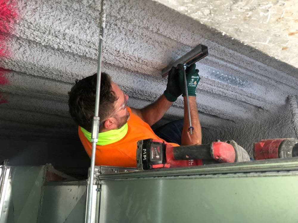 Construction worker applying spray foam insulation in a ceiling with tools and safety gear.