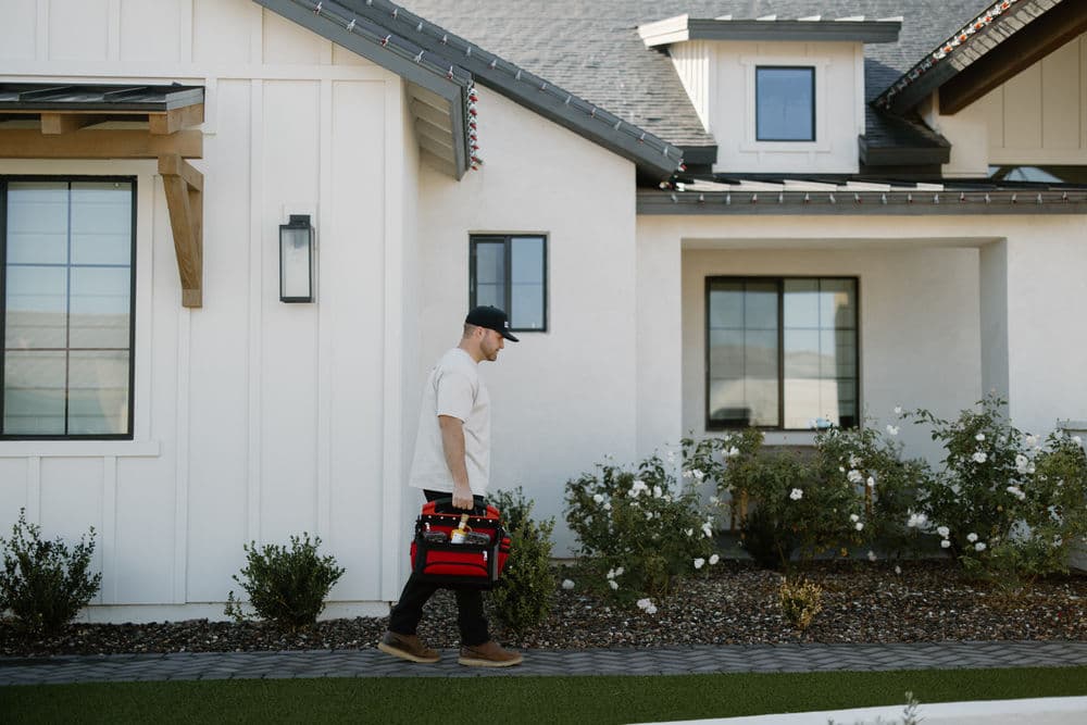 Man carrying a toolbox walking past a modern home with landscaped flowers.